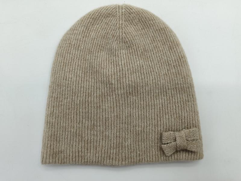 Beżowa prążkowana czapka beanie z małą dzianinową kokardką z boku, na białym tle.