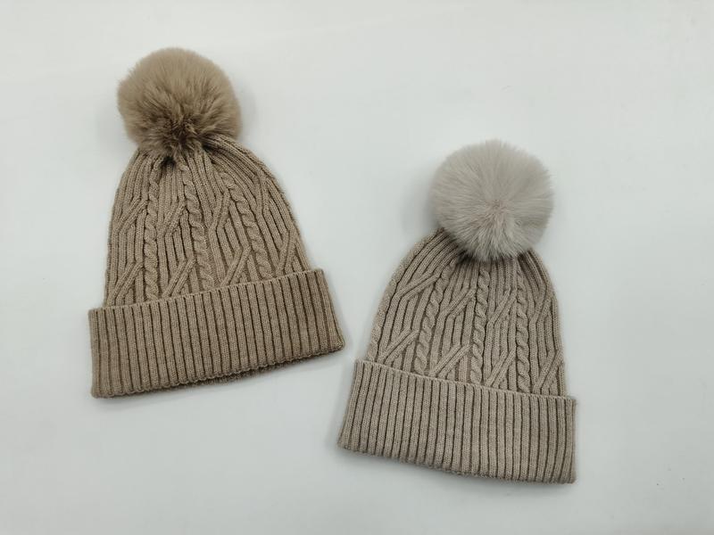 Dwie czapki zimowe cable knit z pomponami z futra syntetycznego w kolorze taupe i jasnoszarym na białym tle.