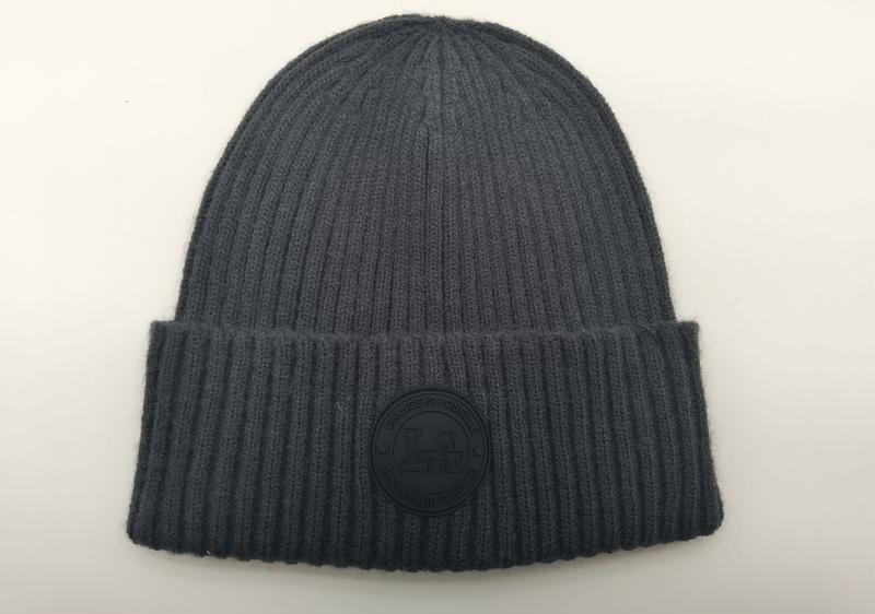 Szara czapka beanie z prążkowanej dzianiny z mankietem i okrągłą gumową naszywką z logo, na białym tle.