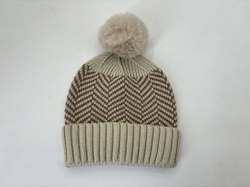Kremowa czapka beanie ze wzorem chevron w kolorze brązowym i puszystym pomponem z sztucznego futra na białym tle.