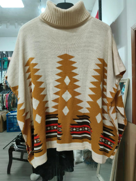 Kremowy sweter poncho z golfem, wzór aztecki w odcieniach musztardy i czerwieni, oversize, na wieszaku.