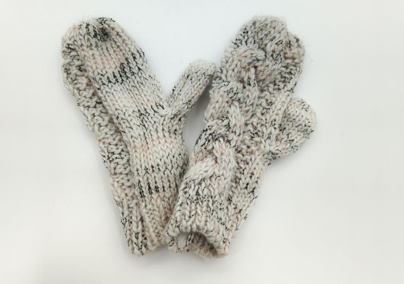 Kremowe rękawiczki chunky knit z czarnymi plamkami i kciukiem, ułożone płasko na białym tle.
