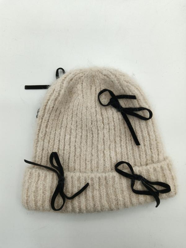 Kremowa czapka beanie z grubym prążkowanym splotem i czarnymi aksamitnymi kokardkami na białym tle.