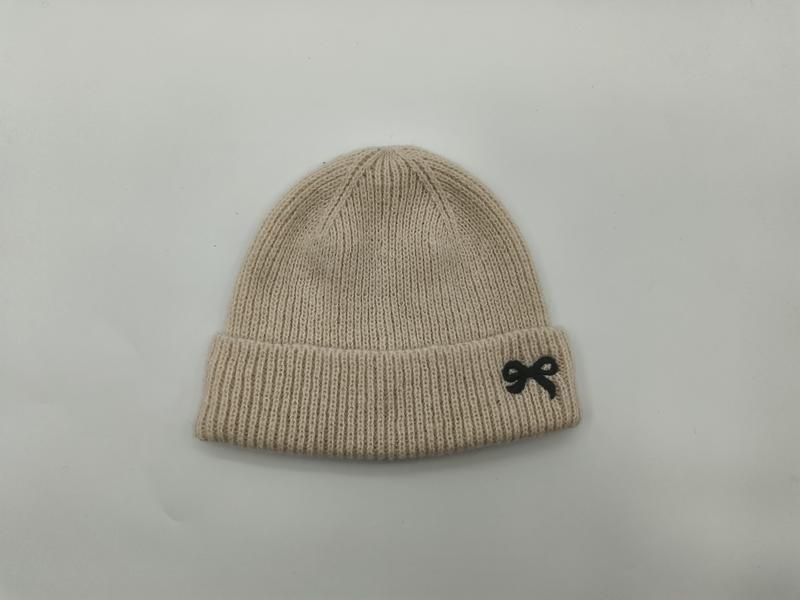 Kremowa prążkowana czapka beanie z czarnym haftem w kokardę na podwiniętym mankiecie, na białym tle.
