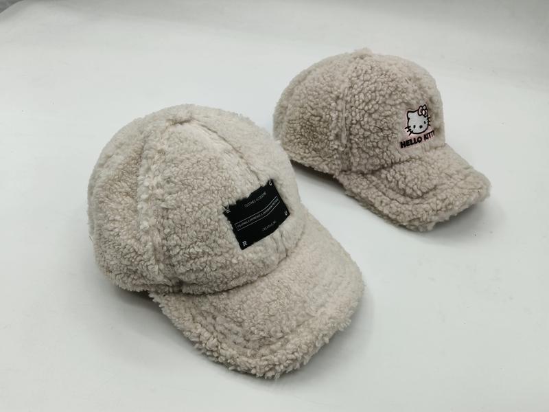 Dwa kremowe czapki baseball z materiału sherpa – jedna z czarną naszywką, druga z haftem Hello Kitty na białym tle.