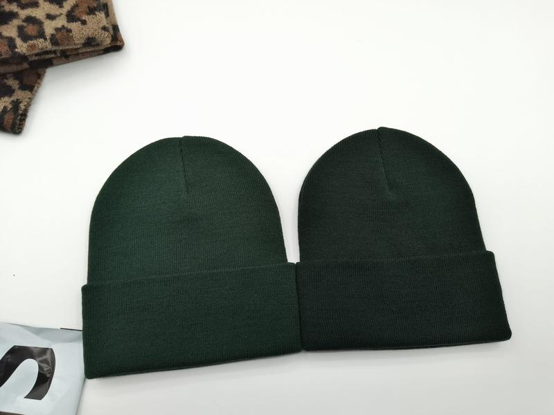 Dwa ciemnozielone prążkowane czapki beanie z mankietem na białym tle obok tkaniny w cętki leoparda.