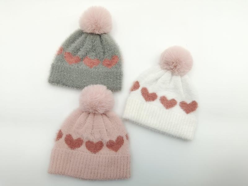 Trzy puszyste czapki beanie z dzianiny ze wzorem w serca i pomponem ze sztucznego futra w kolorach szarym, różowym i białym.