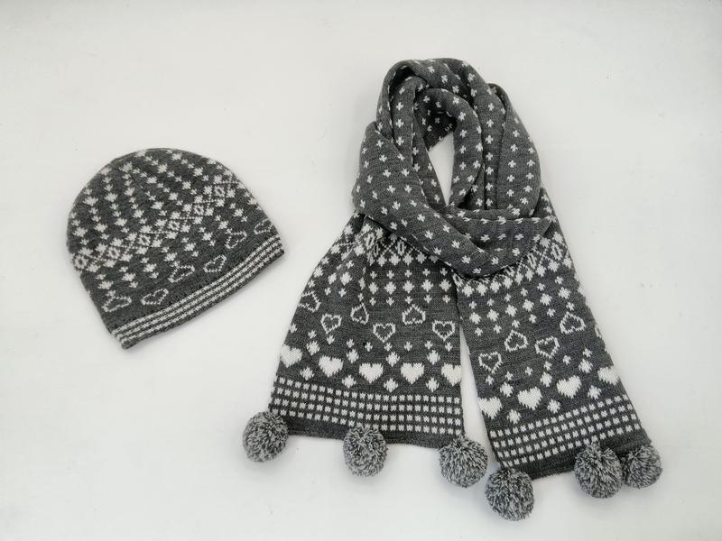 Szaro-biały komplet: czapka beanie i szalik z dzianiny Fair Isle we wzór serc z pomponami na białym tle.