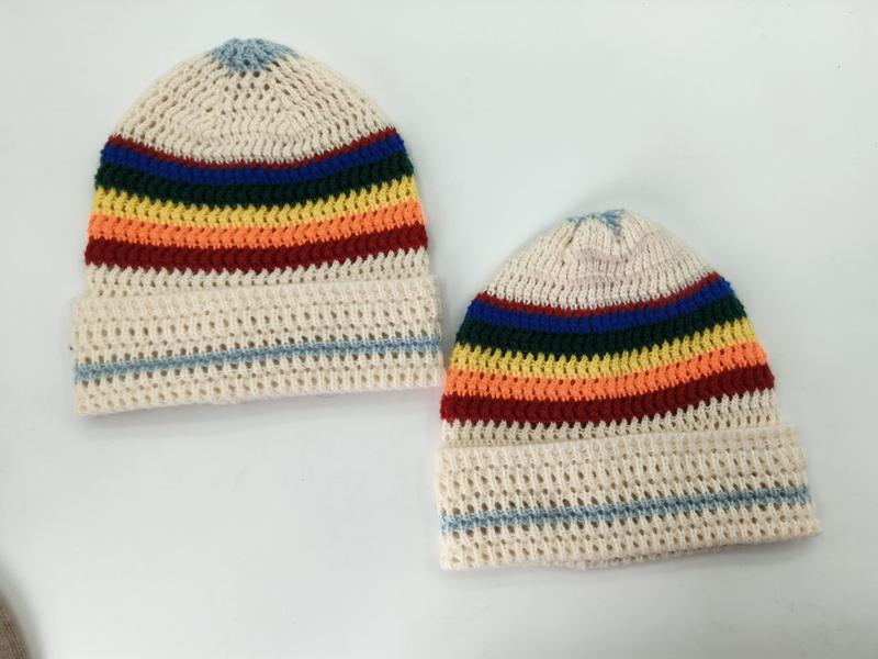 Dwie kremowe czapki beanie na szydełku z tęczowymi paskami w kolorach niebieskim, zielonym, żółtym, pomarańczowym i czerwonym.