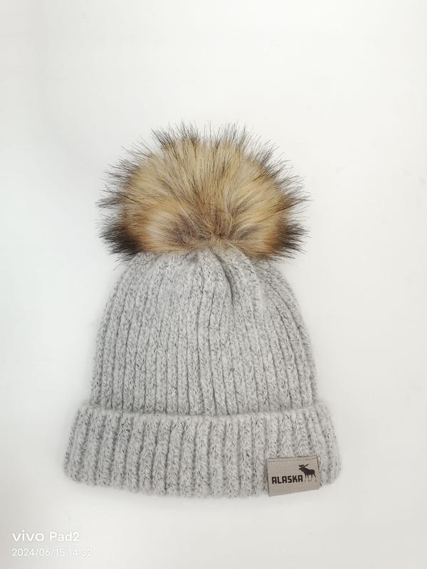 Jasnoszara gruba czapka zimowa beanie w prążki z dużym pomponem ze sztucznego futra na białym tle.