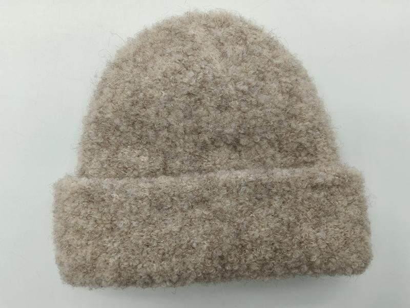 Beżowa czapka beanie z wywijanym mankietem, wykonana z puszystej przędzy boucle w kolorze owsianki, na białym tle.