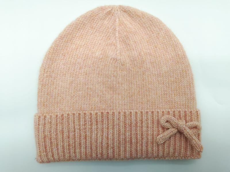 Brzoskwiniowa czapka beanie z prążkowanym mankietem i dekoracyjną kokardką z dzianiny na jasnym tle.