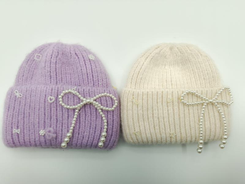 Dwie czapki beanie w kolorze lawendy i kremowym z perłowymi kokardkami i haftowanymi motywami na białym tle.