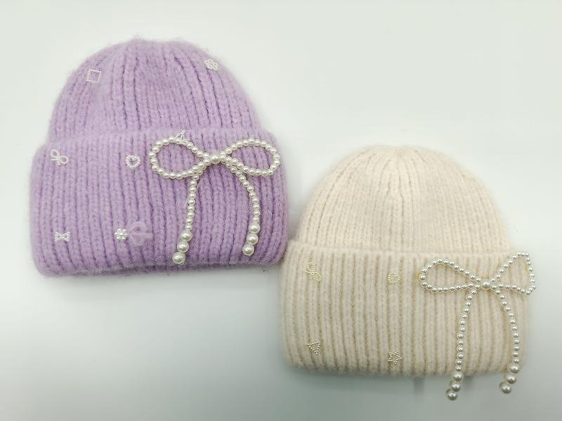 Dwie grube czapki beanie z dzianiny w lawendzie i kremie z perłowymi kokardami i haftowanymi ikonkami.