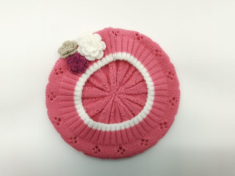 Różowy beret z dzianiny z białym paskiem, koronkowym ażurem i trzema szydełkowymi kwiatkami w bieli, mauve i beżu.
