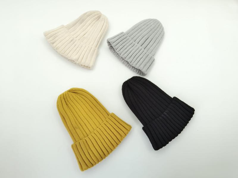 Cztery prążkowane czapki beanie w kolorach: kremowym, jasnoszarym, musztardowym i czarnym, ułożone na białym tle.