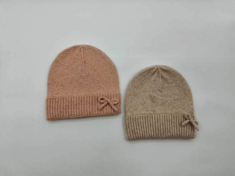 Dwie miękkie czapki beanie z prążkowanym mankietem i kokardką w kolorze różowym i beżowym, na białym tle.