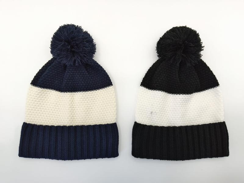 Dwie dzianinowe czapki beanie z pomponem: granatowo-kremowa w paski (lewa) i czarno-kremowa w paski (prawa), białe tło.